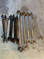 Photo showing 8 wrenches grouped on a concrete surface, three are black open-end wrenches and five are silver combination wrenches.