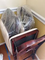 Three folding dining chairs stacked together, partially covered in plastic, showing dark wood frames and white upholstered seats.