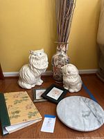 Photo showing two white ceramic cat statues, tall floral porcelain vase, two small framed floral artworks, floral photo album, and round white marble trivet.