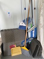 Photo showing 7 household cleaning tools including two brooms with blue bristles, a yellow broom, two dustpans (yellow and red), a blue mop with white microfiber cloth, and a white floor duster, set against stacked dark grey Craftsman interlocking floor tiles and black trim.
