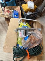 View of cardboard box holding multiple electrical items including a green extension cord, plastic bags with supplies, a yellow container, some packaged light bulbs and electrical components.