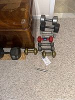 Various hand weights arranged on the floor next to a wooden chest, includes multiple pairs and a single dumbbell