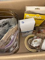 Box with various tapes including Scotch brand packing tape, tape rolls, and other packaging materials.