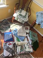 Varied pile of holiday linens including folded tablecloths, runners, and placemats on floor and chairs under window light.