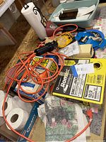 Photo showing a mix of household tools and supplies including an orange electrical cord, yellow work light, boxes, paper towels, and other small items.