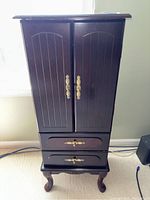 Front closed view of wooden dark wood jewelry armoire with brass-toned hardware and two lower drawers.