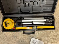 Laser level with tripod and manual inside the open black carrying case on carpet.