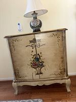 Front view of painted wooden cabinet with floral basket motif and table lamp on top