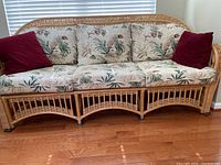 Front view of the rattan sofa showcasing the floral cushions and burgundy throw pillows on hardwood floor.