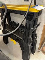 Two stacked folding plastic sawhorses viewed from angle showing yellow tops and black folding legs, stored in a basement area