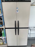 Front view of the full plastic storage cabinet showing all four doors with two upper and two lower doors, black horizontal division, and beige colored door panels with long black handles.