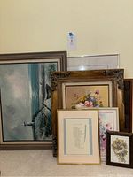 Full view of grouped framed artworks leaning against wall on carpet, showing various sizes and styles including largest painting with lighthouse and floral needlework.