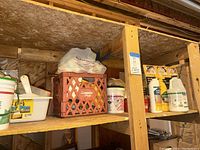 Wooden shelving unit with assorted buckets, bottles of tile grout sealer/removal, and plumbing materials stored on basement shelves.