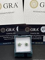 Pair of green moissanite earrings in velvet presentation box with two GRA certificates positioned behind them.