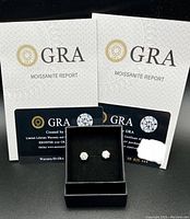 Photo shows the pair of white Moissanite stud earrings in a black box with two GRA Moissanite Report certificates and warranty cards behind them.