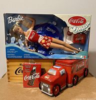Photo showing the unopened 1999 Coca Cola Splash Barbie doll, the wooden Coca Cola box, Coca Cola deck of playing cards, and the red Coca Cola delivery truck tin with wheels.