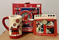 Photo of Coca Cola Christmas themed collectibles with coffee mug, cookie plate and tumbler box, and collectible tin with playing cards.
