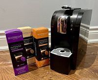 Side angle view of Starbucks Verismo coffee maker with three boxes of Bestpresso K-Fee pods (Intenso, Deciso, Cremoso) placed in front on wooden floor.
