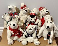 Photo showing ten Coca-Cola branded polar bear plush toys in various hats, scarves, and accessories, grouped together on a flat surface against a white wall