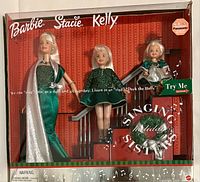 Front view of boxed Barbie, Stacie, and Kelly dolls in green holiday outfits with stairwell accessory in background. Packaging shows brand names and singing sisters logo.