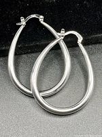 Pair of large, smooth, teardrop-shaped silver hoop earrings with hinged clasp closure, shown on black background.