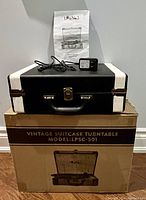 Closed black and beige vintage suitcase turntable placed on top of original box with instruction manual and power cable on top.