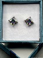 Top view of the pair of square-cut mystic topaz stud earrings in a light blue box with white padded interior, showing iridescent stones and silver prongs.
