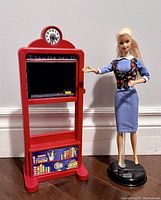 Full view of Barbie teacher doll standing next to red blackboard stand with clock and chalk ledge.