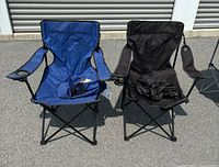Two camping folding chairs placed on pavement, one blue and one black. Both have armrests and black metal frames. The carrying cases are visible on the chairs.