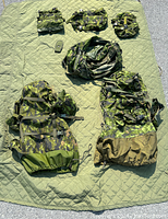 Seven military carrying bags of various sizes and an olive green quilted camping blanket laid out on the ground.