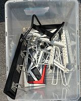 Clear top-down view of a plastic bin filled with mixed brackets and hooks of various materials and colors including black plastic, white wood, and metal parts.