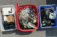 Photo of three plastic bins filled with assorted electronic wires, power packs, and cables placed outdoors on concrete surface.