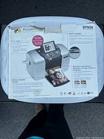 Photo of the Epson PictureMate printer box showing the printer image and product details on the back panel, highlighting features like easy photo printing from memory cards and lab-quality results.