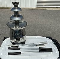 Photo of the stainless steel chocolate fondue fountain with three tiers and six fondue forks lying beside it on a white surface outdoors.