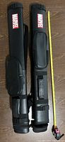 Two Marvel branded black pool cue carrying cases laid parallel on the floor with a tape measure indicating length.