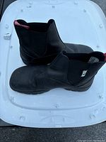 Pair of used black safety boots placed on white surface showing side and top profiles.