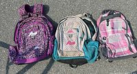 Photo showing three school backpacks including two Roots brand backpacks and one pink plaid backpack.