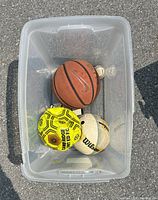 Container holding one basketball, one volleyball, and one soccer ball on pavement.