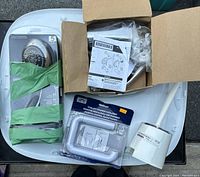 Photo showing boxed showerhead, shower plumbing kit, toilet paper holder in packaging, and white toilet brush with holder on a white surface.