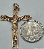 Photo showing front view of gold crucifix pendant next to a 10 cent coin for size comparison, highlighting detailed figure of Christ and filigree cross.