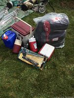 Photo showing coolers, water jugs, small propane tank, sleeping bags wrapped in plastic, and boxed stove on grass.