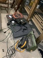 Photo showing flat dolly, hanging trouble light with orange extension cord, knee pads, 2 space heaters (green and black/yellow), all piled on or near the dolly in basement setting.