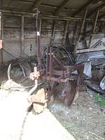 Side front angle view of the vintage tractor plow showing the large moldboard and framing in a barn setting, highlighting its heavily rusted metal condition and base blade.