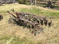Front and side view of rusty older farm disc plow with multiple disc blades and metal frame on grass.