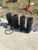 All 4 Lasko black portable tower heaters lined up outdoors on concrete surface showing front and top views, cords wrapped at base, grill front, control buttons on top.