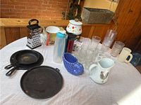Overview photo showing cast iron skillets, fry pan, various glasses, grater, pitchers, and water bottles on round table with white cloth.