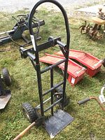 Moving cart/dolly standing vertically on grass, showing black metal frame, arched handle, and base platform with wear and some paint chipping.