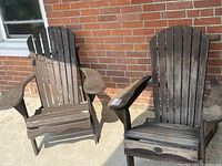 Two wooden Muskoka chairs side by side on concrete patio near brick wall, showing overall wear and weathering on wood surfaces.