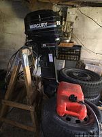 Full view of outboard motor on wooden stand with fuel tank and spare tires in background