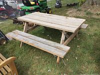Full side view of weathered wooden picnic table with attached benches on grassy yard with garden equipment visible in background.